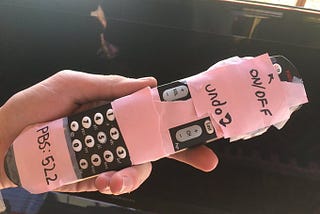 A TV remote with the entire front except the channel and volume buttons covered by pink masking tape.