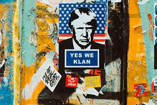 A colorful picture of a mural covered with posters, one showing Donald Trump’s face on an american flag background. Underneath his face is a written sign “Yes we Klan”