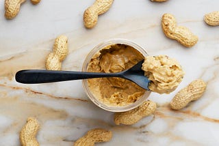 looking down on a table which has scattered peanuts in their shells, at the center is an open jar of chunky peanut butter with a spoonful of the peanut butter balanced on top of the jar