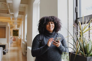 A black woman with natural hair smiles as she holds her phone in an office hallway.