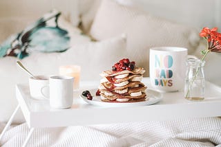 Stock image of a fancy breakfast try on a bed or sofa featuring a stack of pancakes with fruit and several coffee mugs.