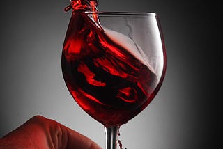 A full glass of red wine, held at the stem by a person’s fingertips, is sloshed over the edge.