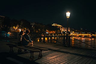 Lover sits on a bridge in Paris with an illuminated lamp post & stunning night scene.