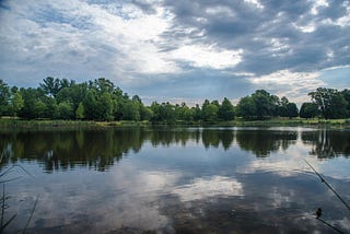 A lake surrounded by trees reflects a blue sky and clouds.