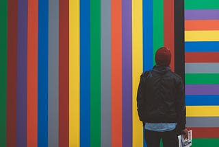 A man holding a newspaper stares at a wall painted with brightly colored vertical stripes