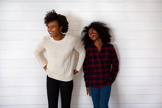 Two darker-skinned women laughing hard. They are wearing sweaters and dark pants and one is leaning against a wall.
