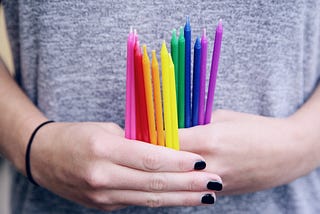 A pair of female hands with black nail polish holds multicolored candles in front of a gray t-shirt.