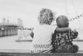 Two children sitting on a bench looking at a large body of water, the older child’s arm around the younger one