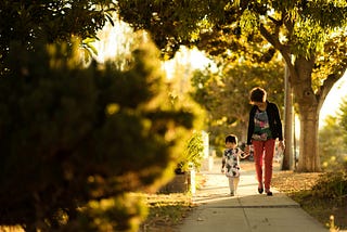 A mother walks her 2 year old daughter down a sidewalk in a sunny afternoon shaded by trees.