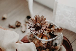 A round glass bowl full of dried flowers and petals sitting on a wooden desk, with the edges of a white lace curtain in the back.