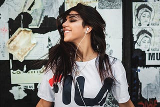 Woman with dark hair smiling and looking off to the side with a white t-shirt on, headphones in, and some designs and doodles pinned on a wall behind her