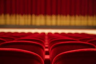 A theater. Perspective from the audience. Rows of empty red velvet chairs and in the distance, a red curtain with gold trim.