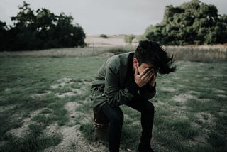 Photo of a person sitting in a field with hands over their face, looking slumped over and upset as if they have been crying or are crying.