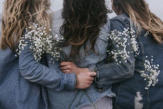 3 ladies wearing denim jackets, ladies on the left and right are holding white flowers with arms behind the lady in the middle