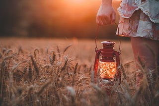 A woman holds a lit lantern in a field of wheat at sunset.
