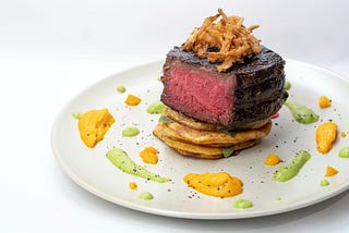 A medium rare steak fillet cut to show the inside. It is beautifully plated with dabs of orange and yellow sauce around a white plate. Delicious-looking fried onions are piled atop the meat. Yum!