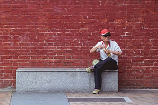 A man sits alone in front of bright red wall, opening a bottle.