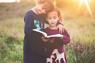 Two little girls sharing the joy of a book while standing in a sunlit field.