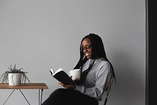 A black woman with braids is sitting and reading a black book with a white up in her hand. Next to her is a table with a plant on top.