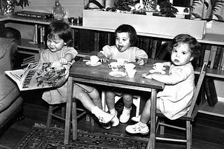 A black and white photograph of three toddlers sitting around a small table with a play tea set spread between them