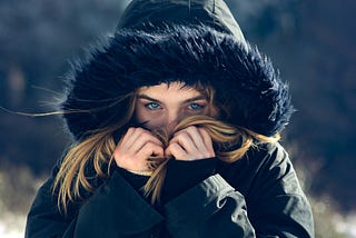 Young woman in a winter jacket with a fur-lined hood standing in a snowy landscape. She peers out with visible blue eyes, her hands covering her mouth and nose, partially obscuring her face. The cold, wintry background contrasts with the warmth of her clothing.