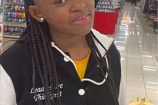 Nya Jingles is a 15 year old teen. She is an African American girl. She is seen in a grocery store with her hair in braids, a yellow shirt on and a black and white letterman jacket.