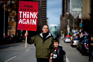 A man and child holding up a sign that says “Make America Think Again”