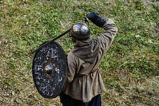 A medieval/Viking cosplayer is seen from above, standing alone in a muddy, grassy field. No face is visible. The individual’s back is to the viewer. They wear a metal helmet without horns, carry a dull metallic shield and hold their dull metal sword across their chest. They are dressed in a brownish-grey tunic top with a hood, the material is coarse. Their pants are billowed and navy blue. Their boots are brown leather and with canvas uppers.