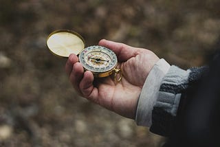 A person’s hand holds an old, gold compass, against a backdrop of autumn leaves and layered sleeves.