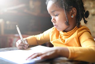 Black girl writing with a pen on paper. She is wearing a yellow shirt and has braids in her hair. She appears to be concentrating.