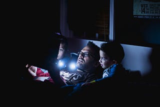 A father and his two young children are sitting in bed at night, illuminated by the light of a tablet or book they are looking at together. The father, wearing glasses, is in the middle, with a child on each side, all appearing engaged and absorbed in the story. The room is dark, with the soft glow of the screen lighting their faces, creating a cozy and intimate atmosphere.