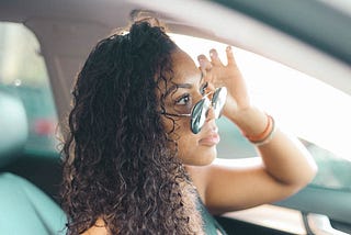 Lady with long curly hair looking over sunglasses while driving.