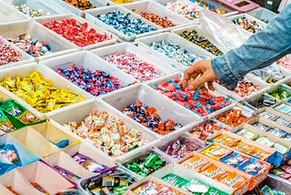 A colorful assortment of candies displayed in open plastic bins at a store, with a person reaching to grab a piece. The bins include various types of individually wrapped sweets and packs of gum.