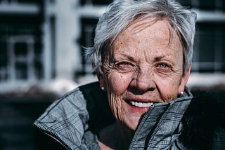 Elderly woman standing outside with a coat on zipped up to the neck.