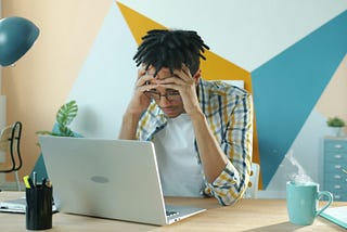 A person sits at a desk holding their head in frustration while looking at a laptop screen. The workspace includes a teal mug with steam rising from it, pens, and a desk lamp. The background wall features bold geometric shapes in teal, mustard yellow, and white, adding a modern feel to the scene. The image conveys stress or difficulty concentrating while working. Photo by Vitaly Gariev on Unsplash