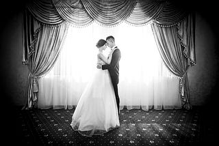 A monochrome image showing man a and woman in wedding outfits embracing in front of a large window in what could be a hotel function room.