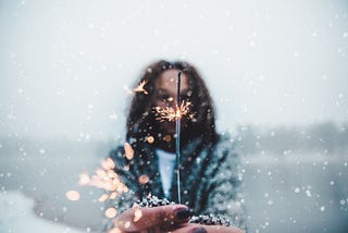 A woman stands outdoors in the snow. She holds a sparkler, which is lit and sparkling in the foreground.