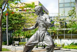 A statue of Bruce Lee, shirtless, in fighting stance, outside a glass building
