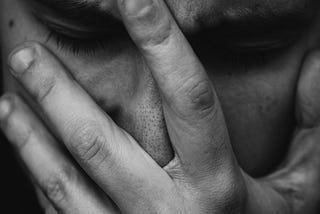 Black and white close up photograph of a man in discomfort, one hand on his face in a feeble attempt to stiffle his sobs.