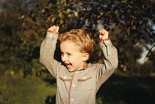 Excited small boy with brown hair and a big smile with his hands above his head