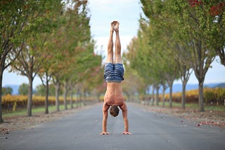 A man performing a handstand in the middle of a tree-lined road, surrounded by autumn foliage. He is shirtless and wearing shorts, with vibrant greenery and a distant landscape visible in the background, creating a serene and balanced composition.
