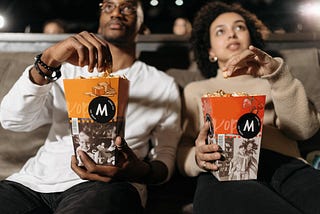 A couple is watching a movie in a theater. We see that each has their own large popcorn.
