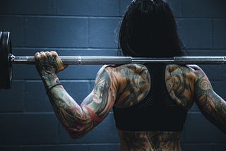 Image of tattooed girl’s back holding weighted barbell on neck
