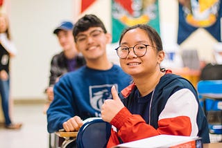 Close up of two students sitting at their desks in a classroom. The girl students has a thumbs-up sign. Both kids look happy.