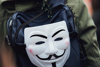 A man with a hooded jacket walking away from the camera. He is wearing a backpack, on which a Guy Fawkes mask hangs, looking directly at the camera.