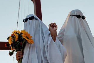 A couple holding hands with flowers and putting on white blankets as ghost costumes with sunglasses on