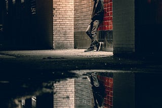 A young man stands in a dark alleyway, reflected in a puddle on the ground. His face his hidden by his hoodie, he leans against a brick wall alone.
