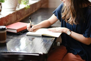 Picture of a woman sitting at a black counter wearing a blue shirt and brown pants writing in a journal, facing a window.