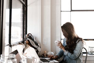 Girl wit long brown hard in a jean jacket holds a cup of coffee while scrolling on her laptop at a counter by the window in a coffee shop. Her backpack is on the counter along with other cups and another laptop.