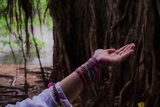 A woman’s hand with bright bracelets is palm up near a large tree.